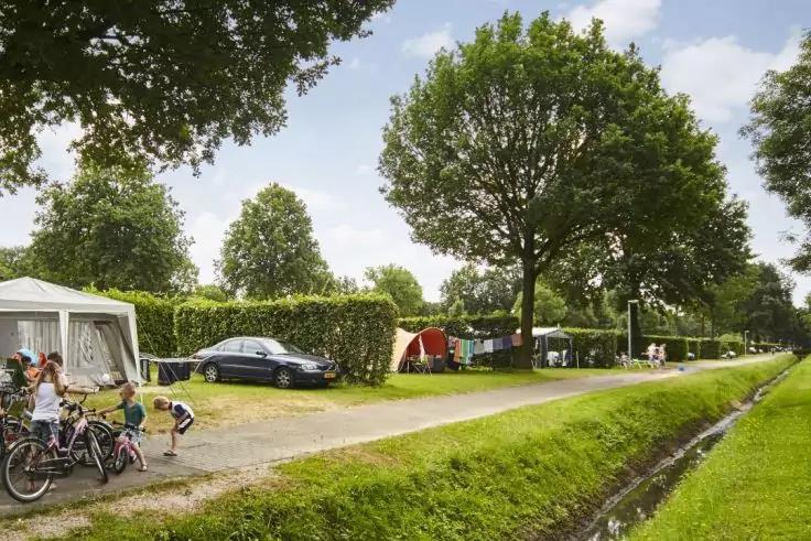 Campingplatz mit Rasen, schmalem Kanal rechts, Kiesweg, B&auml;umen und Hecken, wei&szlig;em Vorzelt links, rotem Zelt, dunklem Auto und Kindern mit Fahrr&auml;dern links