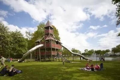 Gro&szlig;er h&ouml;lzerner Spielturm mit zwei Metallr&ouml;hrenrutschen auf Rasen, Personen auf Decken, B&auml;ume, See im Hintergrund, bew&ouml;lkter Himmel