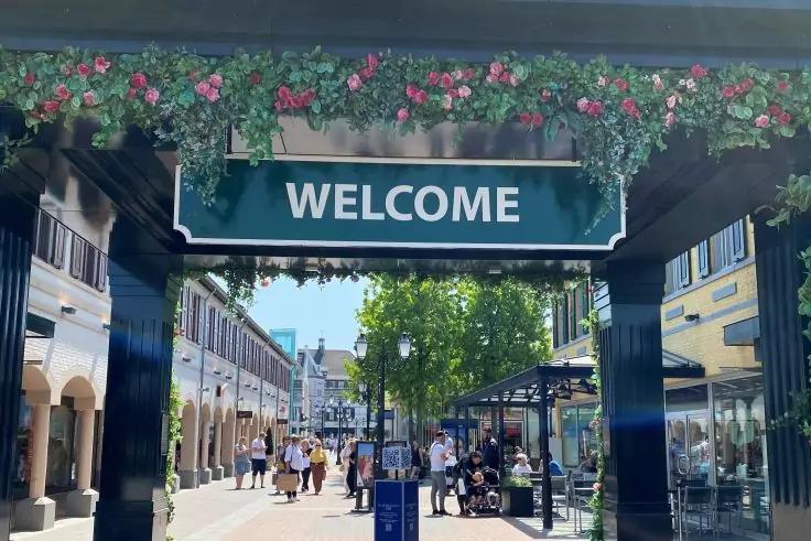 Gr&uuml;nes Schild mit wei&szlig;er Aufschrift 'WELCOME' h&auml;ngt unter einer mit Blumen geschm&uuml;ckten &Uuml;berdachung; gepflasterter Durchgang mit Gesch&auml;ften, B&auml;umen im Hintergrund und mehreren Personen