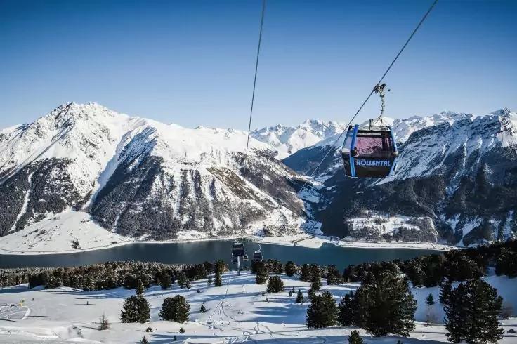 Gondel einer Seilbahn &uuml;ber verschneiter H&uuml;gellandschaft mit vereinzelten Tannen, See und schneebedeckten Bergen unter klarem blauem Himmel