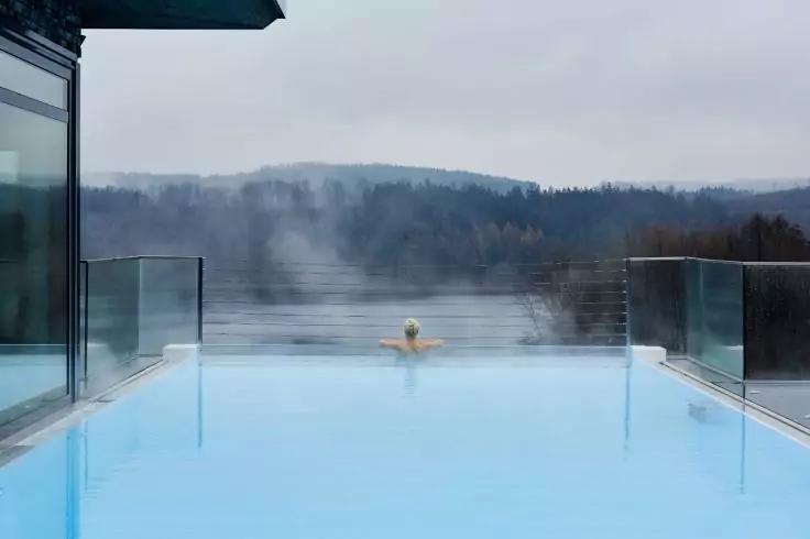 Person im Au&szlig;en-Infinitypool mit R&uuml;cken zur Kamera, lehnt am Beckenrand; Dampf &uuml;ber dem Wasser; Glasgel&auml;nder und Geb&auml;udeecke links; neblige, bewaldete H&uuml;gel im Hintergrund