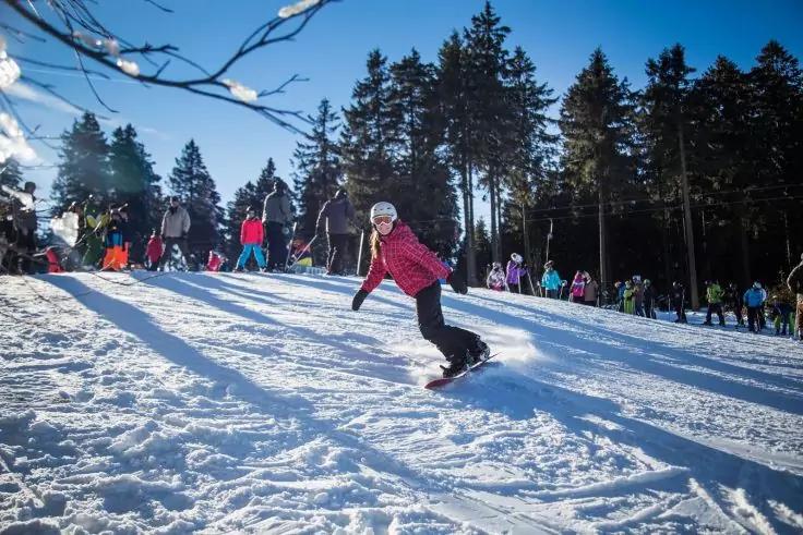 Person auf Snowboard f&auml;hrt einen verschneiten Hang hinab, im Hintergrund mehrere Menschen und Nadelb&auml;ume unter blauem Himmel