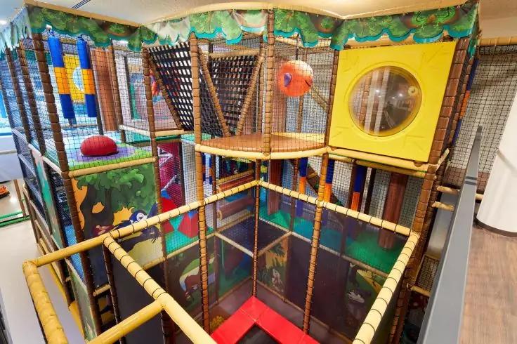 Mehrst&ouml;ckiges Indoor-Spielger&uuml;st mit schwarzem Netz, gelbem quadratischem Panel mit rundem Fenster, orangefarbener Ball, gepolsterten gelben Gel&auml;ndern, bunten Paneelen und roter Bodenmatte