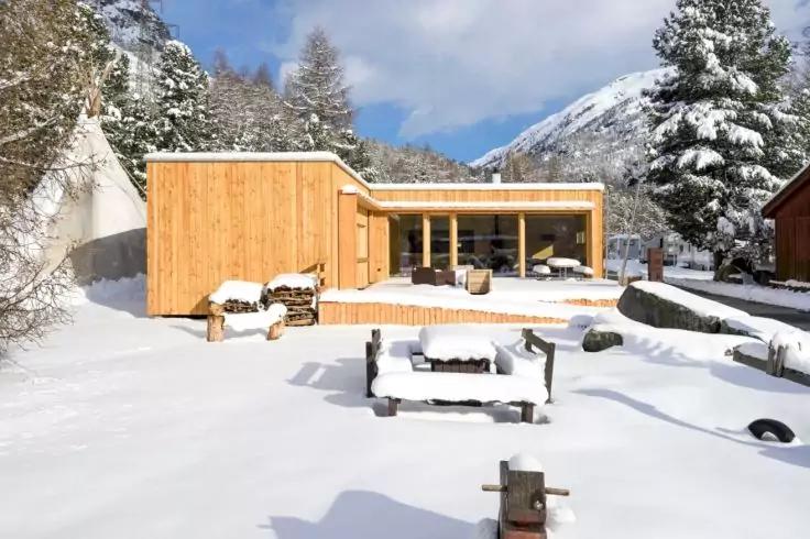 Holzhaus mit gro&szlig;er Glasfront in verschneitem Garten, verschneite B&auml;nke, gestapeltes Brennholz, Tannen und Berge im Hintergrund