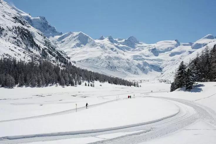Verschneites Tal mit schneebedeckten Bergen im Hintergrund, Nadelb&auml;ume am Hang, geschwungene Spur im Vordergrund und mehrere Personen in der Ferne