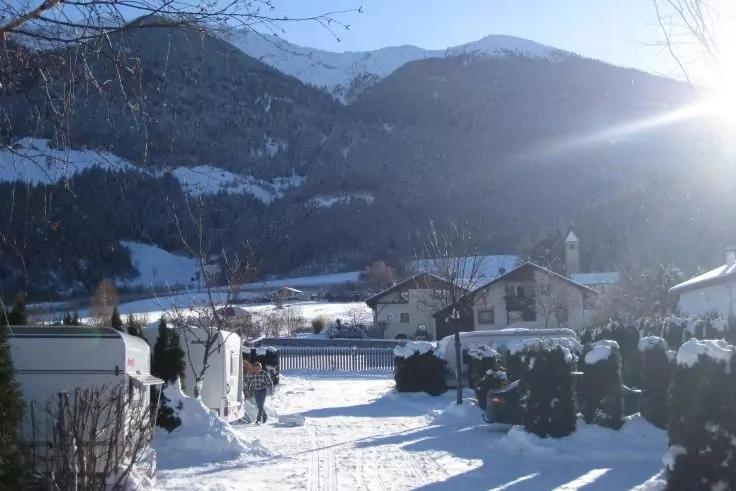 Schneebedeckter Dorfweg mit zwei Personen, Wohnwagen, Hecken, H&auml;usern und Bergen; Sonne rechts oben