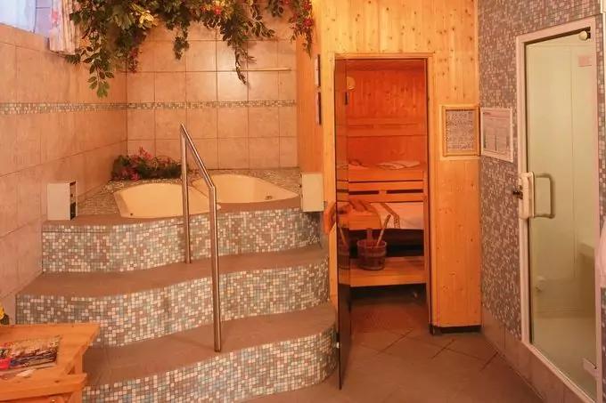 Mosaikverkleideter Whirlpool auf erh&ouml;htem Podest mit Edelstahlgel&auml;nder, daneben ge&ouml;ffnete Holzsaunat&uuml;r mit Holzb&auml;nken und Wasserk&uuml;bel, rechts Glast&uuml;r (Dusche), Kachelw&auml;nde und h&auml;ngende Pflanzen