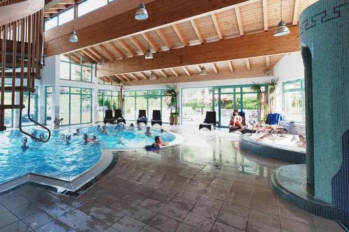 Innenraum eines Schwimmbads mit Becken und Whirlpoolbereich, mehreren Personen im Wasser, Holzdecke mit sichtbaren Balken, gro&szlig;en Glasfenstern und Liegest&uuml;hlen