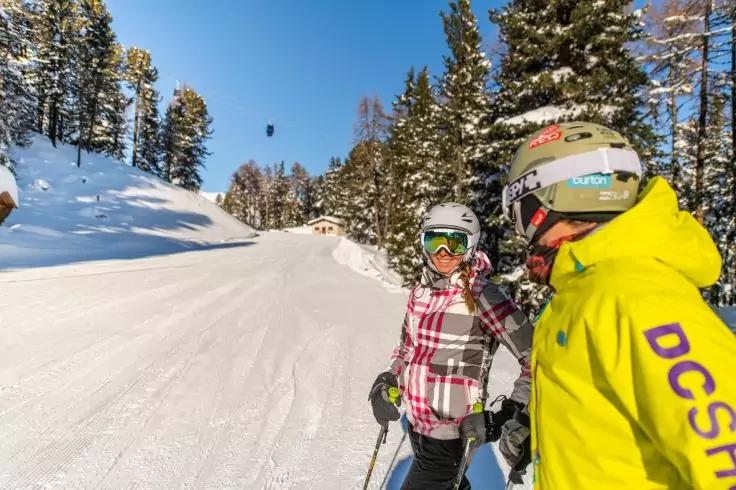 Zwei Personen mit Skihelmen und Skibrillen, eine in gelber Jacke, eine in karierter Jacke, mit Skist&ouml;cken auf einer schneebedeckten Piste; Tannenb&auml;ume und blauer Himmel im Hintergrund