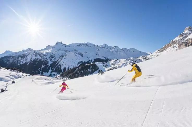 Zwei Skifahrer in gelber und pinker Jacke fahren &uuml;ber verschneite H&auml;nge; sonniger blauer Himmel und schneebedeckte Berge im Hintergrund