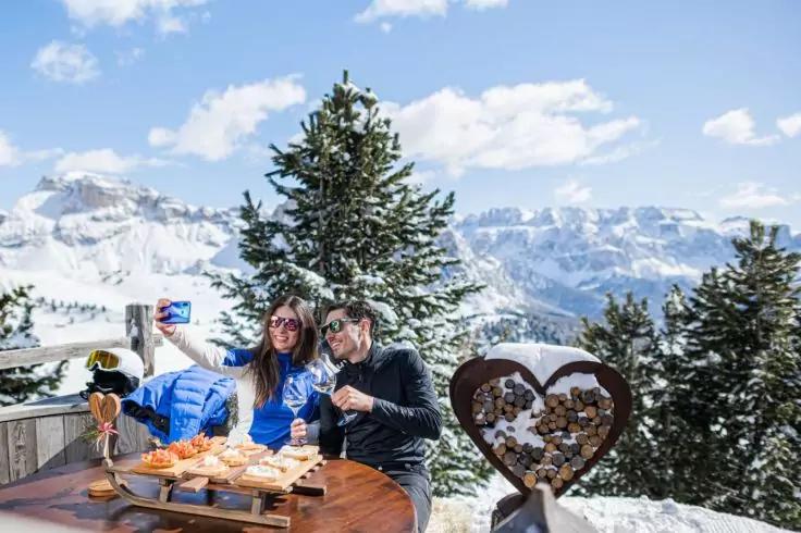 Zwei Personen in Winterjacken und Sonnenbrillen sitzen an einem Holztisch auf einer verschneiten Bergterrasse; eine Person h&auml;lt ein Smartphone f&uuml;r ein Selfie; auf dem Tisch ein Brett mit Geb&auml;ck, Gl&auml;ser und eine herzf&ouml;rmige Holzdekoration; im Hintergr