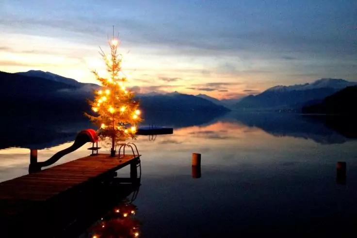 Holzsteg &uuml;ber ruhigem See bei D&auml;mmerung, am Ende ein beleuchteter Tannenbaum, Lichter spiegeln sich im Wasser, Berge im Hintergrund