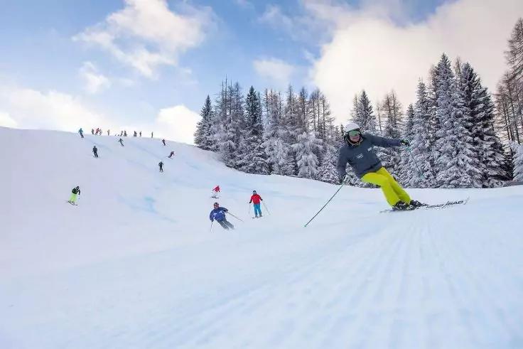 Mehrere Skifahrer auf einer verschneiten Piste; im Vordergrund ein Skifahrer mit gelben Hosen; rechts verschneite Tannen; blauer Himmel mit Wolken