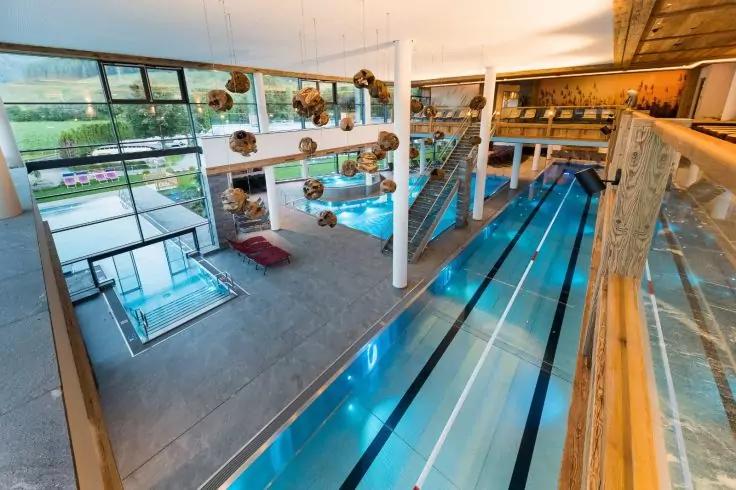 Innenraum eines Hallenbads mit Mehrbahnenbecken, kleineren Becken, gro&szlig;en Panoramafenstern, S&auml;ulen und h&auml;ngenden Kugelleuchten