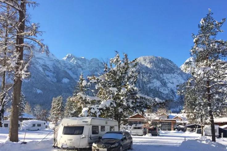 Wohnmobil und Auto im Schnee vor schneebedeckten B&auml;umen, H&auml;usern und Bergen unter blauem Himmel