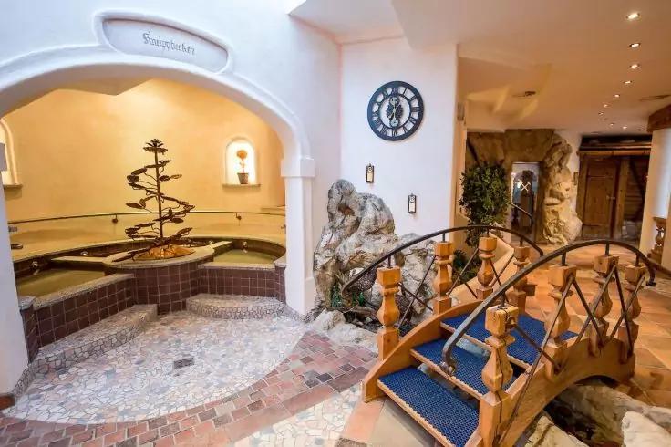 Innenraum mit Mosaikfliesenboden, rundes mehrstufiges Wasserbecken mit kleiner dekorativer Font&auml;ne, h&ouml;lzerne geschwungene Fu&szlig;g&auml;ngerbr&uuml;cke mit blauem Teppich, k&uuml;nstliche Felsformation, runde Wanduhr, Wandnischen mit Leuchten