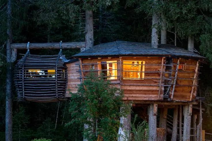 Holz-Baumhaus auf Stelzen zwischen hohen Nadelb&auml;umen, Fenster innen beleuchtet, kleine seitliche Plattform