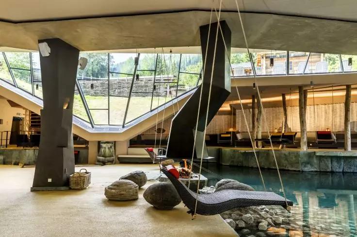 Innenraum mit Wasserbecken, h&auml;ngende Liege an Seilen &uuml;ber dem Wasser, runde Steinkissen auf dem Boden, gro&szlig;e schr&auml;ge Fenster, Sitzbereich mit Sofas und Betonpfeilern
