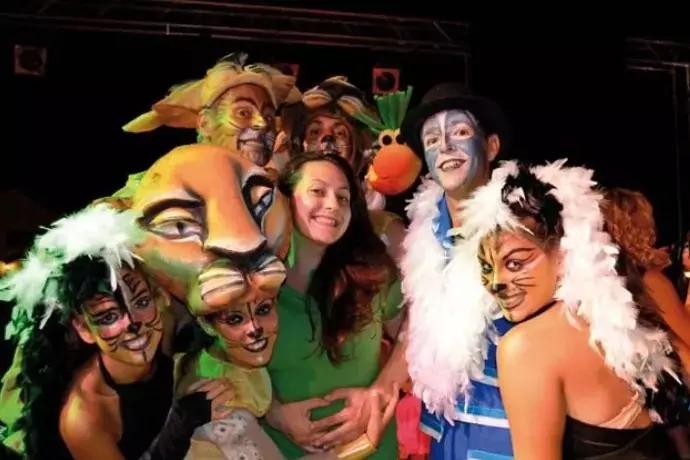 Mehrere Personen mit Tiergesichtsbemalung und Kost&uuml;men um eine Frau ohne Schminke in gr&uuml;ner Bluse; wei&szlig;e Federboas, eine Person im blauen Kost&uuml;m mit schwarzem Hut, eine orangefarbene Handpuppe mit gr&uuml;nem Element, bunte B&uuml;hnenbeleuchtung, dunkler Hint