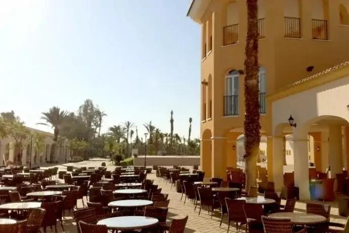 Au&szlig;ensitzbereich mit vielen runden Tischen und St&uuml;hlen auf gepflasterter Terrasse, rechts ein gelb-orangefarbiges Geb&auml;ude mit B&ouml;gen und Balkonen, im Hintergrund Palmen und klarer Himmel