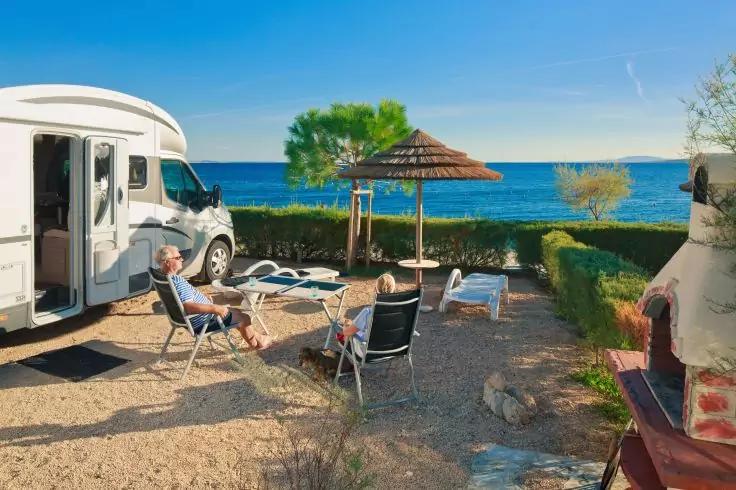 Zwei Personen sitzen auf Campingst&uuml;hlen neben einem Wohnmobil auf einem Platz am Meer; strohgedeckter Sonnenschutz, Liegest&uuml;hle, Hecke, weiteres Wohnmobil, Meer und blauer Himmel sichtbar