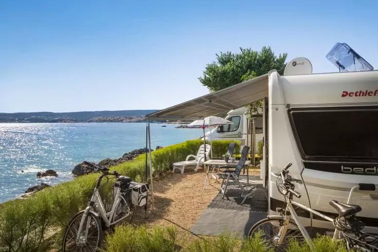 Wei&szlig;er Wohnwagen mit ausgefahrener Markise, zwei Fahrr&auml;der, Klappst&uuml;hle und Tisch am Ufer, Meer und felsige K&uuml;ste unter blauem Himmel