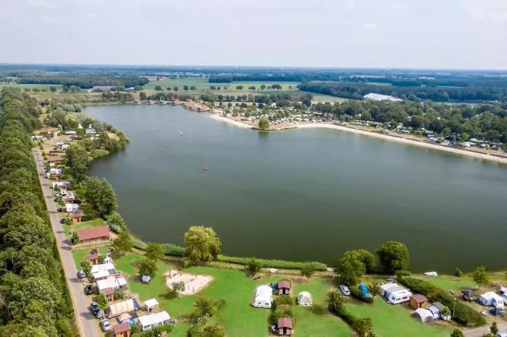 Blick von oben auf einen See mit Ufer und Sandstrand, B&auml;umen, Gr&uuml;nfl&auml;chen, verstreuten Wohnwagen und kleinen H&uuml;tten, einer Stra&szlig;e links und bewaldeter Fl&auml;che im Hintergrund