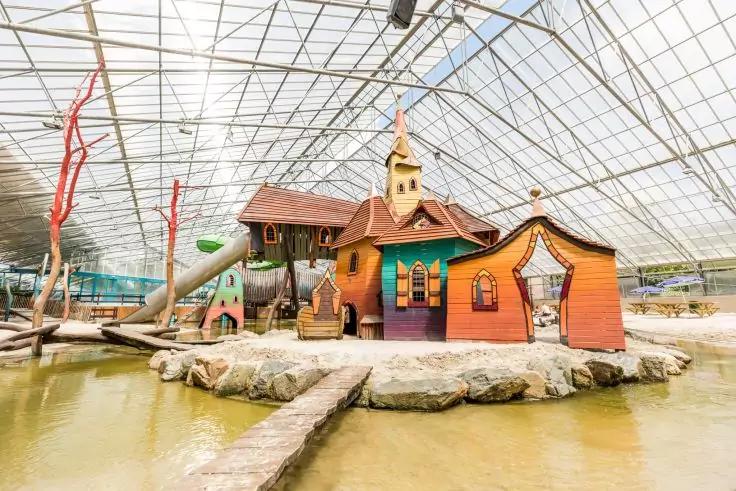 Bunte H&auml;uschen mit T&uuml;rmchen und Rutschen auf einer felsigen Insel in flachem Wasser unter einem gro&szlig;en Glashausdach, mit schmaler Fu&szlig;g&auml;ngerbr&uuml;cke
