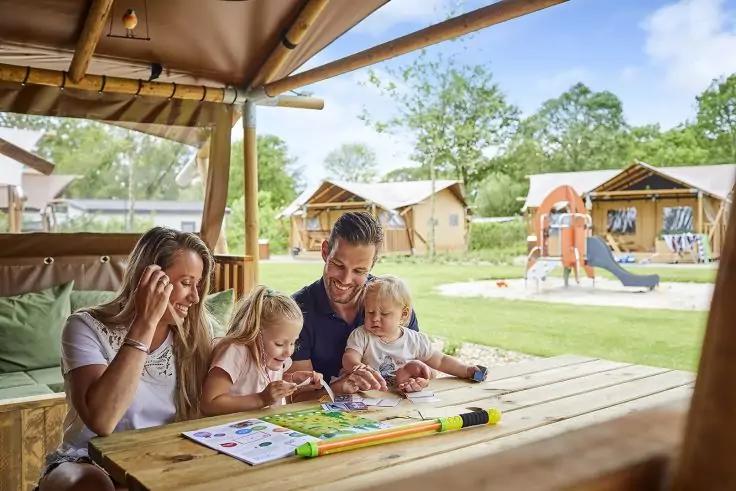 Zwei Erwachsene und zwei Kinder sitzen an einem Holztisch unter einem Dach, die Kinder nutzen ein Malbuch und Zeichenmaterial; im Hintergrund Holzh&auml;user, ein Spielplatz, Rasen, B&auml;ume und blauer Himmel