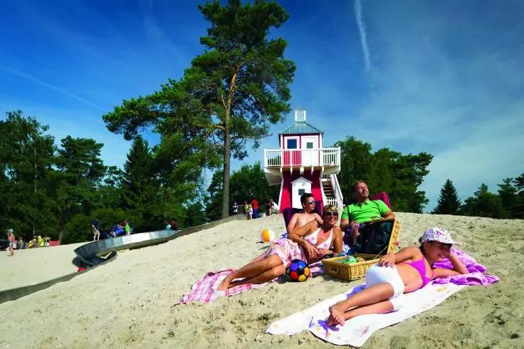 Drei Personen sitzen auf Strandt&uuml;chern im Sand; Korb und Spielzeug neben ihnen; rot-wei&szlig; gestreifte Holzturm im Hintergrund, Kiefern und blauer Himmel mit Kondensstreifen