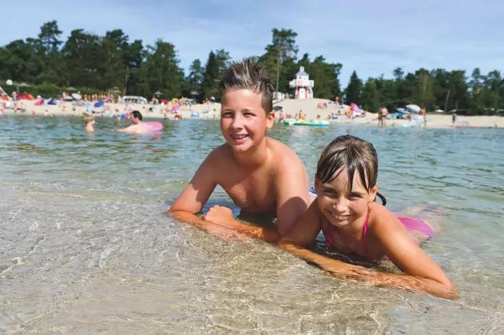 Zwei Kinder liegen bauchw&auml;rts im flachen Meerwasser am Strand, tragen Badebekleidung; im Hintergrund Sandstrand mit Menschen und B&auml;umen, blauer Himmel