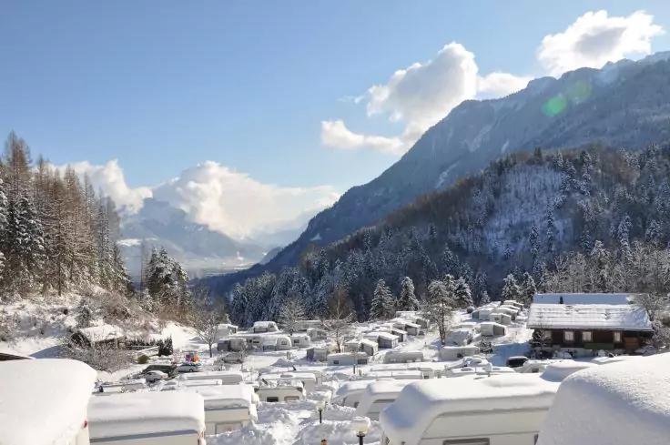 Verschneiter Campingplatz mit vielen schneebedeckten Wohnwagen in einem Tal, Nadelb&auml;ume links, verschneite Berge im Hintergrund und blauer Himmel mit Wolken