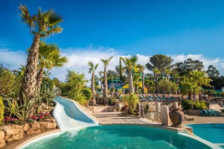 Schwimmbecken mit t&uuml;rkisfarbenem Wasser, kleine wei&szlig;e Wasserrutsche links, Stufen und Felsenrand, Palmen und Pflanzen, blauer Himmel