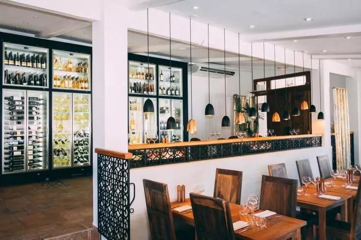 Innenaufnahme eines Restaurants: Bartresen mit h&auml;ngenden Lampen, Glast&uuml;ren-K&uuml;hlschr&auml;nke mit Flaschen, gedeckte Holztische und St&uuml;hle