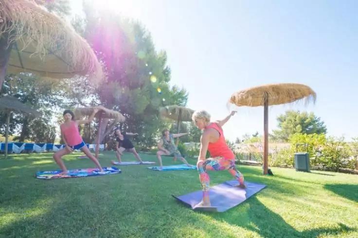 Vier Personen in Sportkleidung auf Yogamatten auf einem Rasen in stehenden Haltungen; Strohh&auml;ute/Sonnenschirme, B&auml;ume und starkes Sonnenlicht im Hintergrund