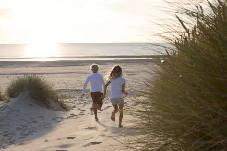 Zwei Kinder in T-Shirt und Shorts rennen &uuml;ber Sandd&uuml;nen zum Strand; D&uuml;nengras links und rechts, Sonnenlicht spiegelt sich auf dem Meer