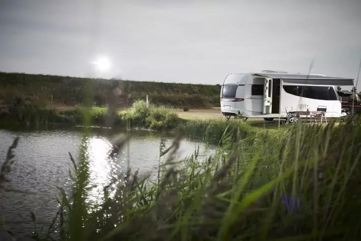 Wohnmobil am Ufer eines Teichs, im Vordergrund Schilf und Wasser mit Sonnenreflexion