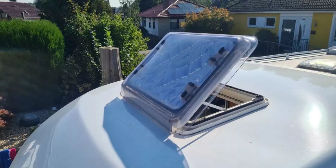 Teilweise ge&ouml;ffnete rechteckige Dachluke mit transparentem Deckel auf wei&szlig; gew&ouml;lbtem Dach, im Hintergrund H&auml;user und Hecke