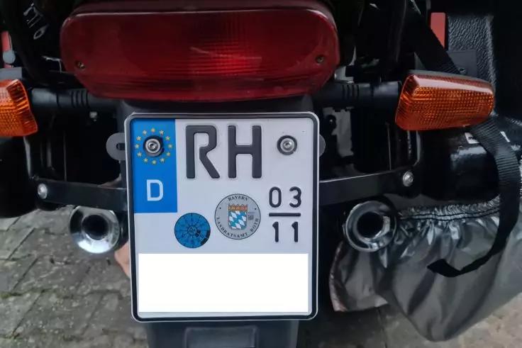 Heck eines Motorrads mit rotem R&uuml;cklicht, zwei orangen Blinkleuchten, zwei Auspuffrohren, rechter Satteltasche und deutschem Kennzeichen mit blauem EU-Streifen, Buchstaben 'RH' und zwei Pr&uuml;fplaketten