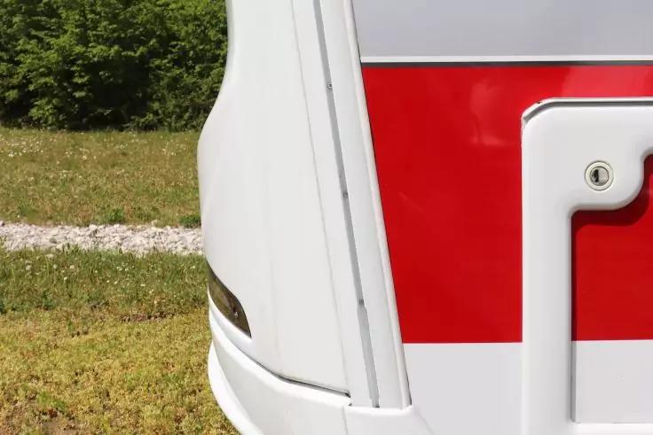 Nahaufnahme der rechten Hinterecke eines wei&szlig;en Fahrzeugs mit rotem horizontalem Streifen; rechteckige Ausbuchtung mit Schraube, kleines gelbes Licht; im Hintergrund Gras, Kiesweg und B&uuml;sche