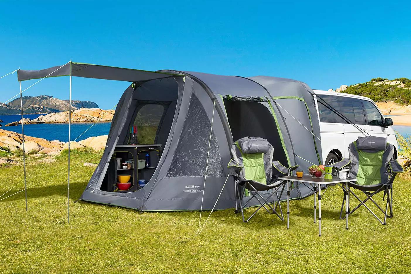 Grau aufgeblasenes Familienzelt mit offenem Eingang und ausgeklapptem Vordach, neben einem wei&szlig;en Wohnmobil auf einer Grasfl&auml;che am Meer; zwei Klappst&uuml;hle und ein kleiner Campingtisch mit Obst davor
