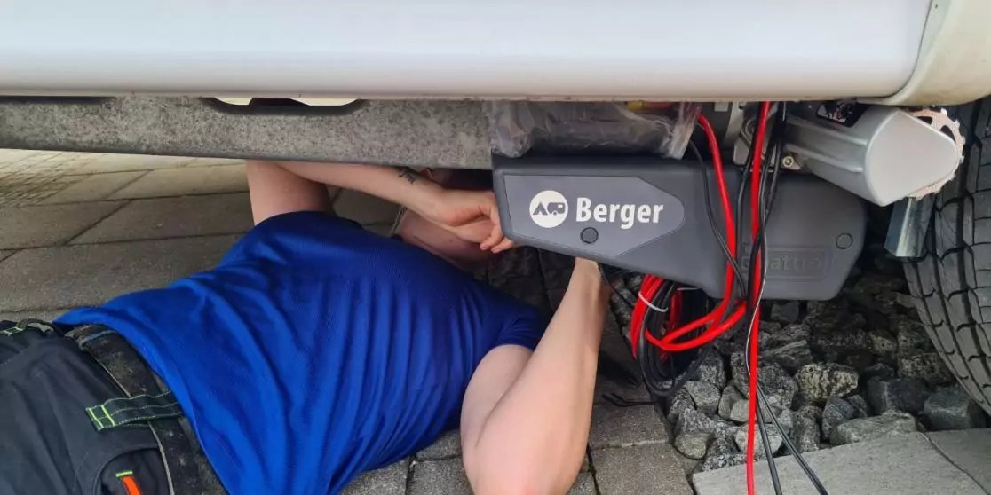 Person lying under a vehicle wearing a blue shirt, hands holding a device labeled 'Berger' with red cables, vehicle tire and paving stones visible
