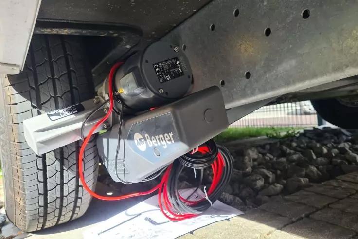 Reifen neben verzinktem Metallrahmen; darunter ein schwarzes Ger&auml;t mit roten und schwarzen Kabeln, ein grauer Stecker, Kies und Pflaster