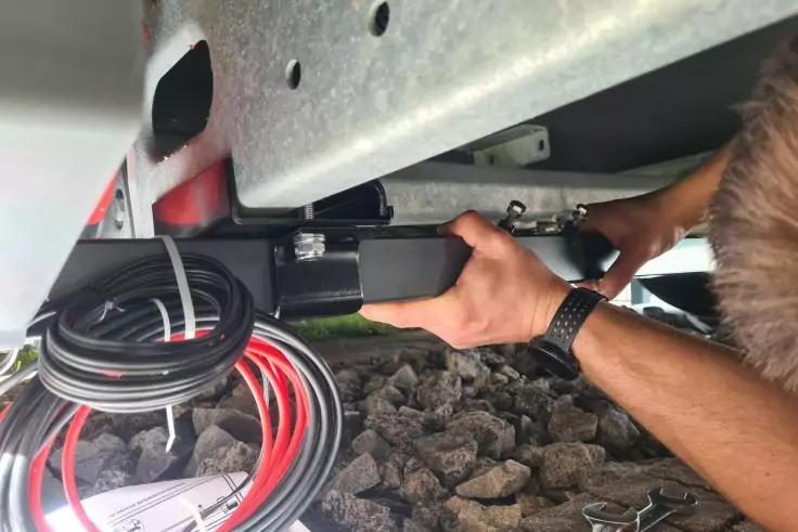 Unterseite eines Metallrahmens; H&auml;nde und Unterarme montieren ein schwarzes rechteckiges Ger&auml;t; aufgewickelte rote und schwarze Kabel liegen auf Kies daneben