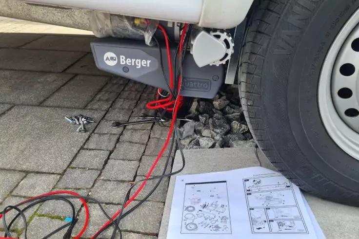 Rechtes Rad eines Fahrzeugs, graues Ger&auml;t mit Aufschrift "Berger" darunter, rote und schwarze Kabel auf Pflaster, ge&ouml;ffnete Anleitungspapiere auf dem Boden, lose Schrauben und Kies