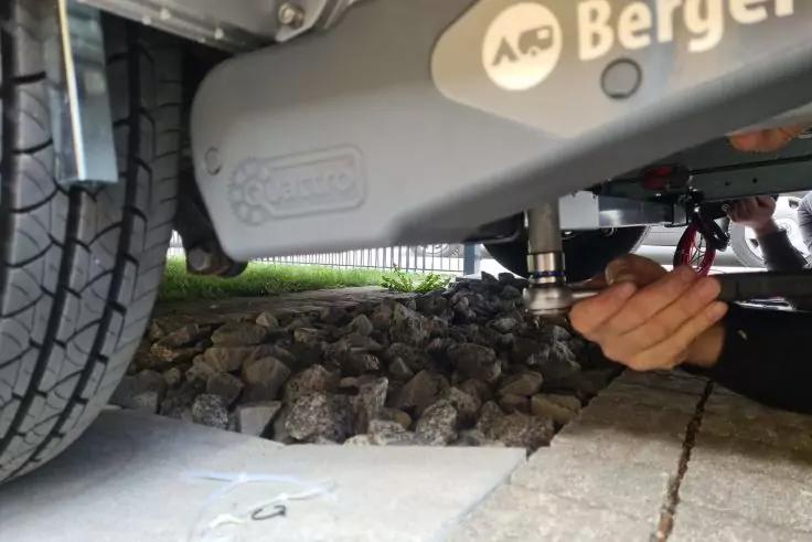 Hand mit Werkzeug unter einer Metallachse mit Schriftzug &bdquo;Berger&ldquo;, links ein Reifen, Boden aus Betonplatten und Kies