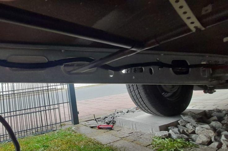 Unterseite eines Fahrzeugs mit Metallrahmen, rechter Reifen, Pflastersteinen, Gras links, Metallzaun im Hintergrund und rotem Wagenheber auf dem Boden