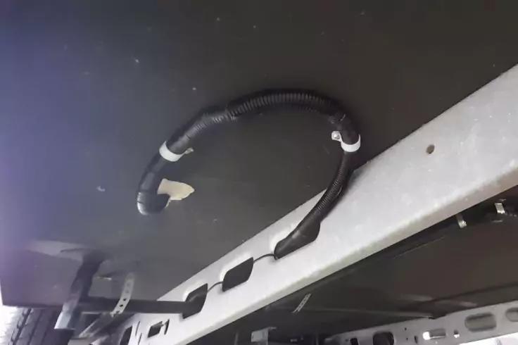 Schwarze, gewellte Kabelschutzleitung, an einer dunklen Platte und einem grauen Metalltr&auml;ger befestigt; wei&szlig;e Klebestreifen und Befestigungsclips sichtbar