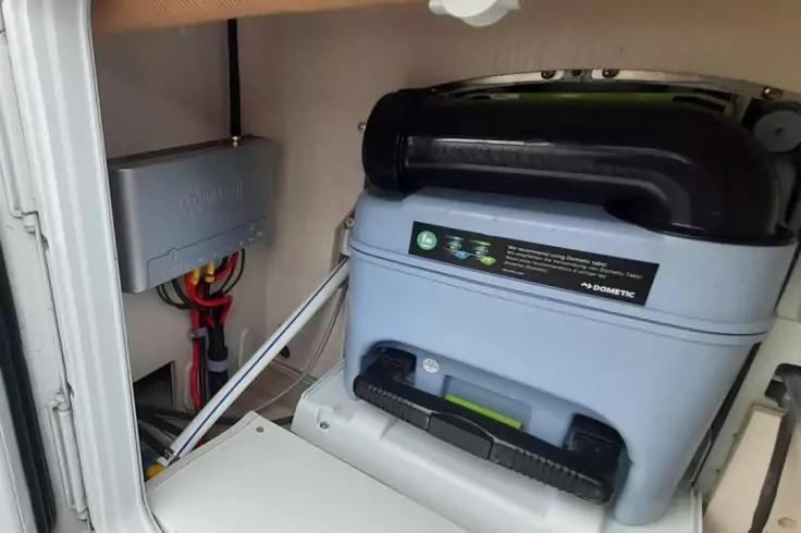 Grauer Kunststoffbeh&auml;lter mit schwarzem Tragegriff in offenem Schrankfach; links eine graue Box mit sichtbaren roten und schwarzen Kabeln