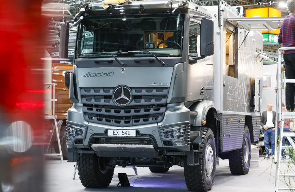 Vorderansicht eines grauen Mercedes-Lastwagens mit Mercedes-Stern und Kennzeichen 'EX 540', unscharfer roter Bereich links, Teile von Personen und eine Leiter rechts, Regale und Deckenbeleuchtung im Hintergrund